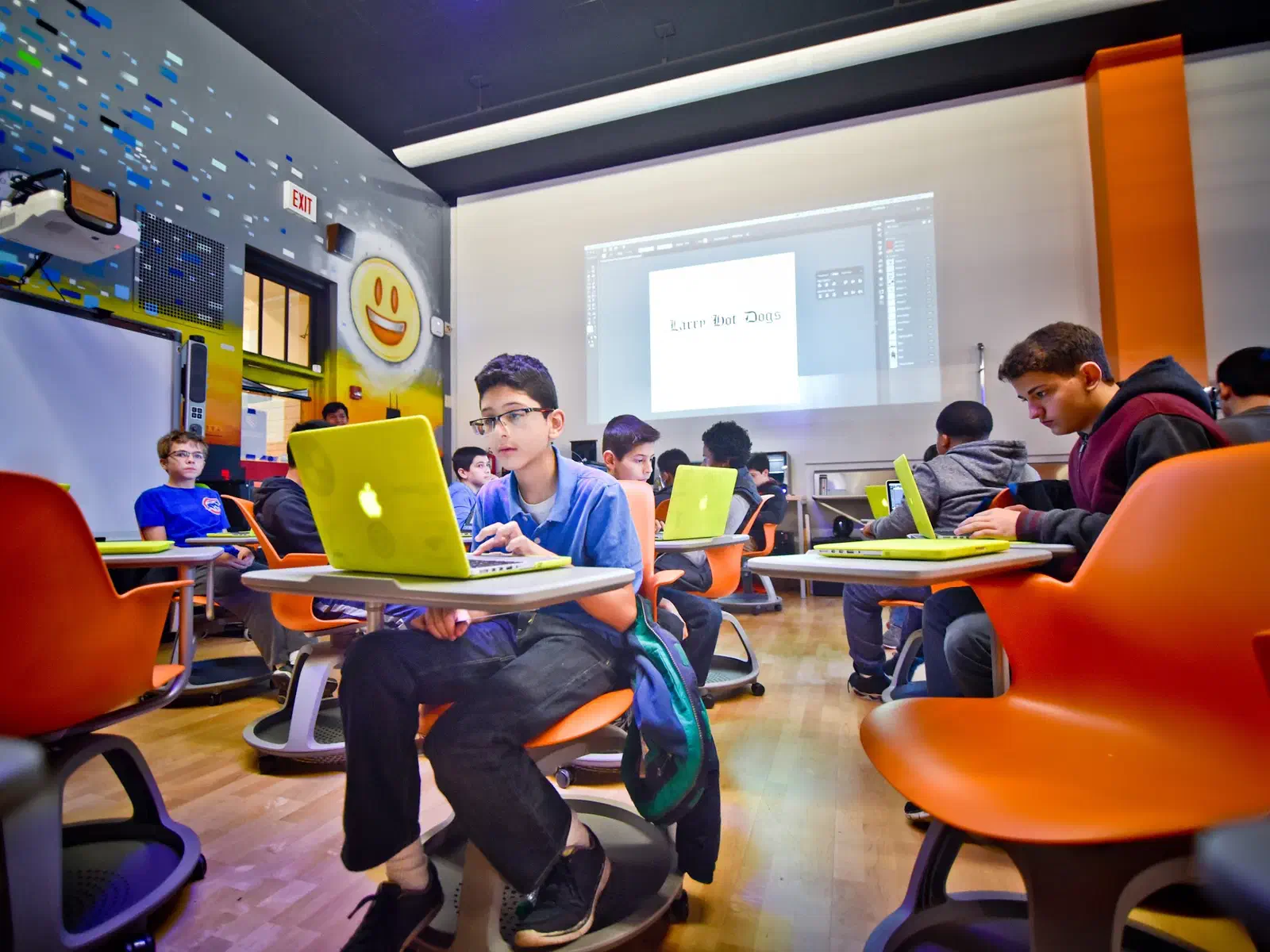 A high school classroom with students designing on laptops at orange desks, a mural on the wall behind them.