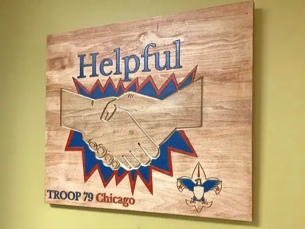 A Scout troop plaque with the word Helpful above two carved hands shaking, painted blue and orange.