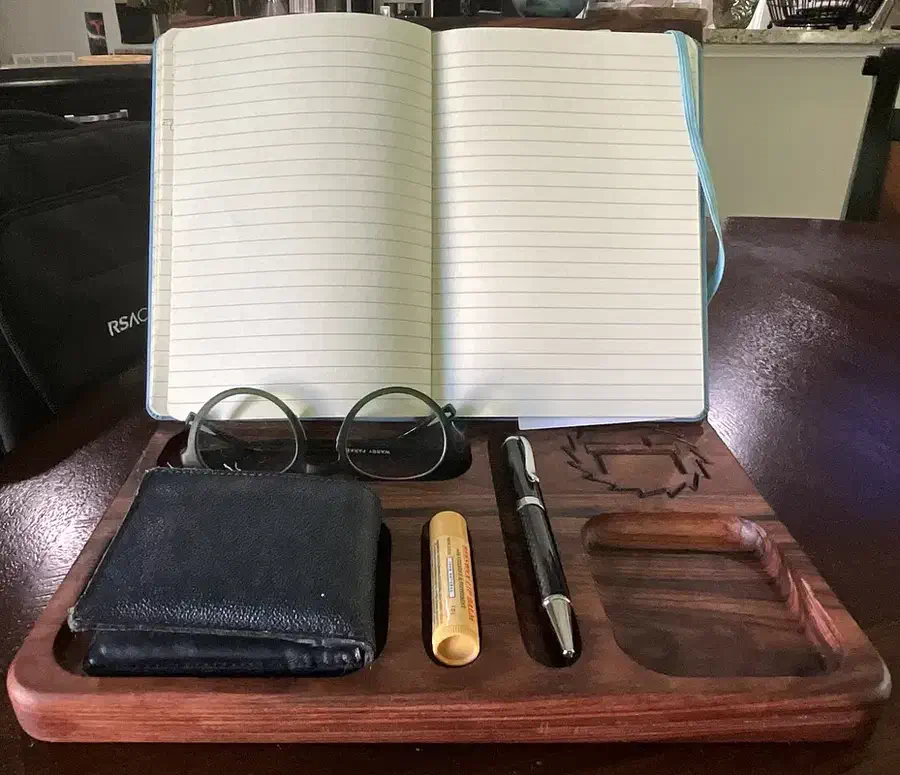 An open notebook on a wooden stand above a tray holding glasses, a wallet and a pen