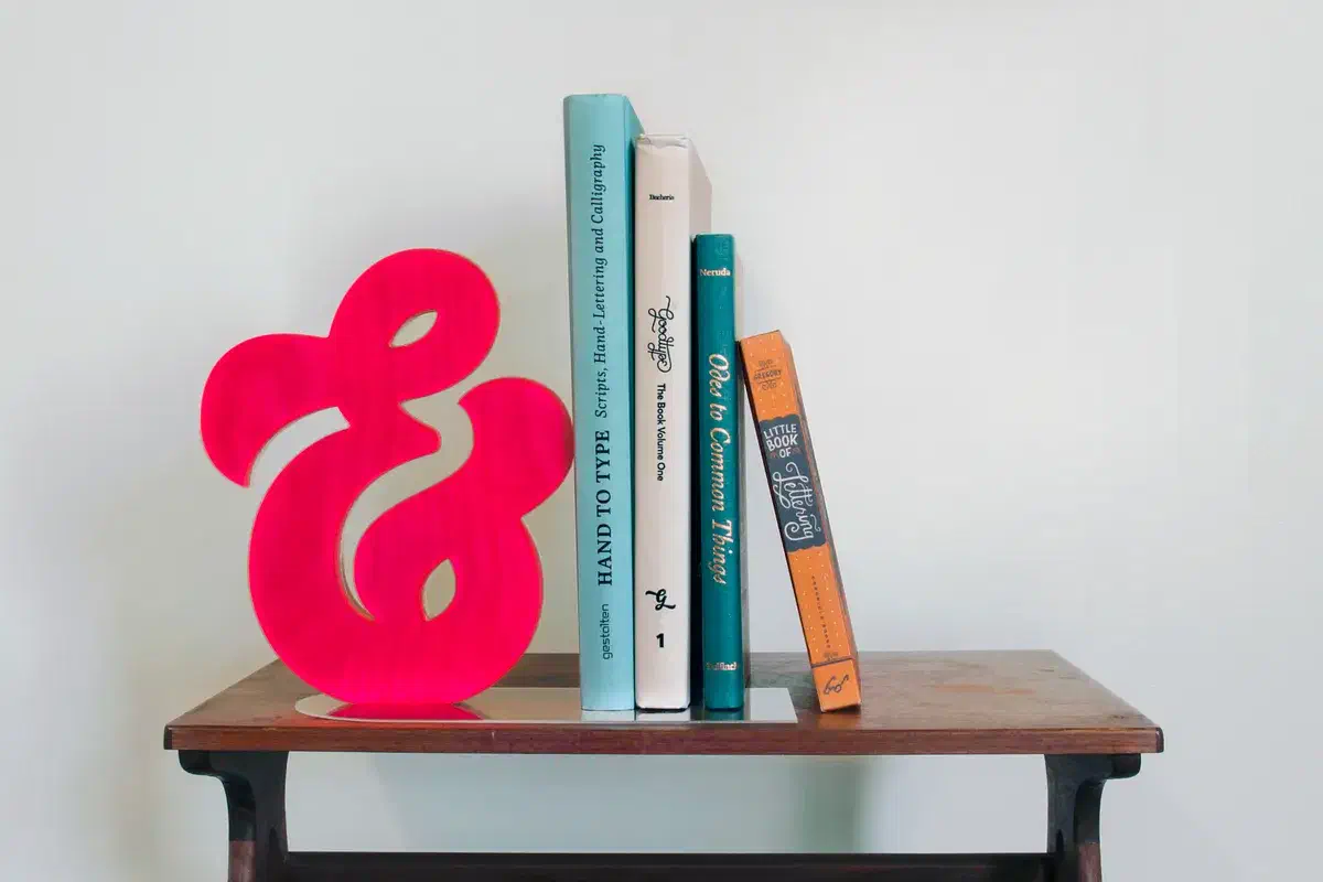 Whole pink acrylic-faced plywood ampersand on a clear base beside four books, against a plain pale wall.