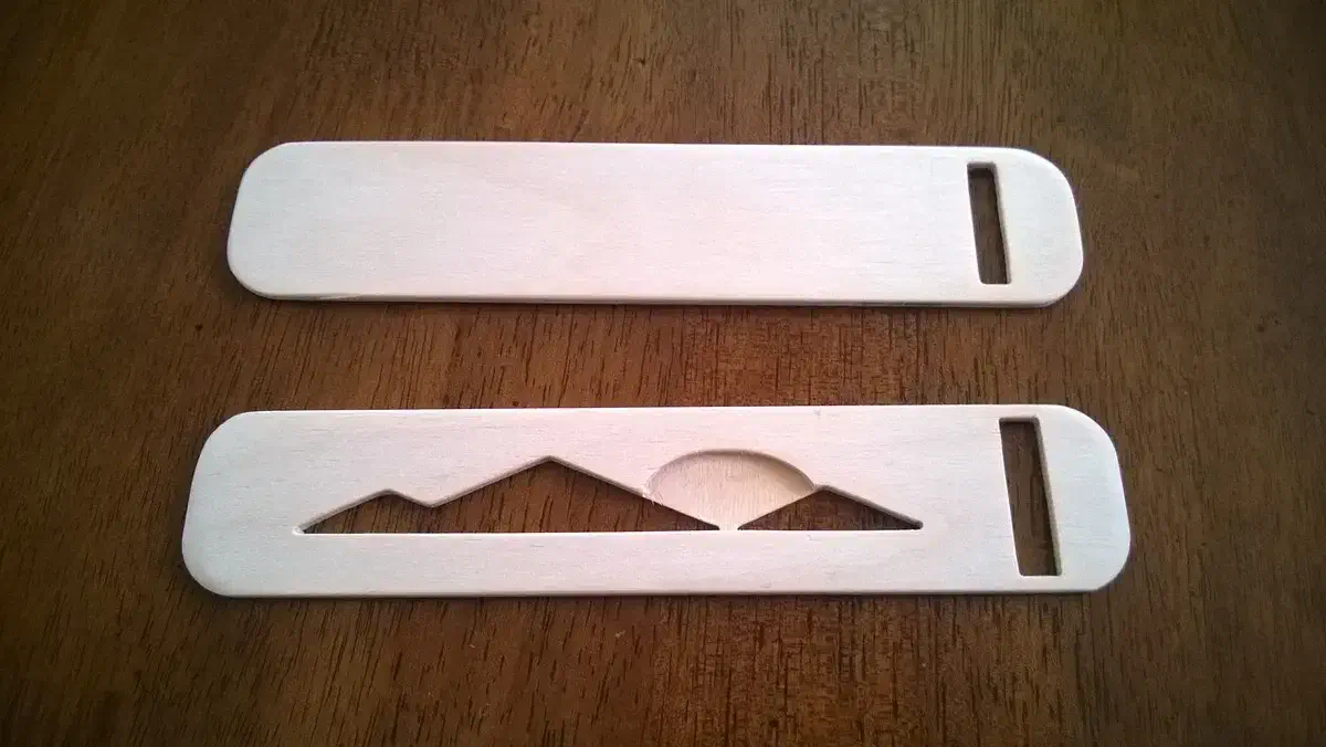 Two complete pale plywood bookmarks on a wooden table, one plain and one with a mountain-and-sun cutout, both with slots at the right end