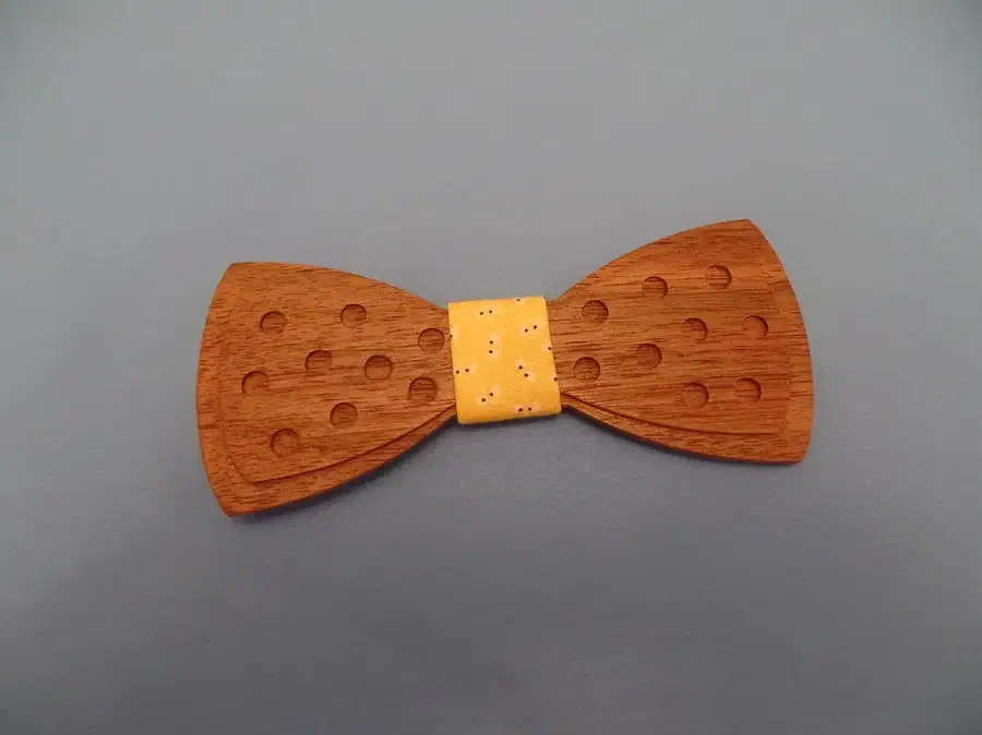 A complete walnut bow tie with recessed dots and a yellow fabric center on an uncluttered gray background.