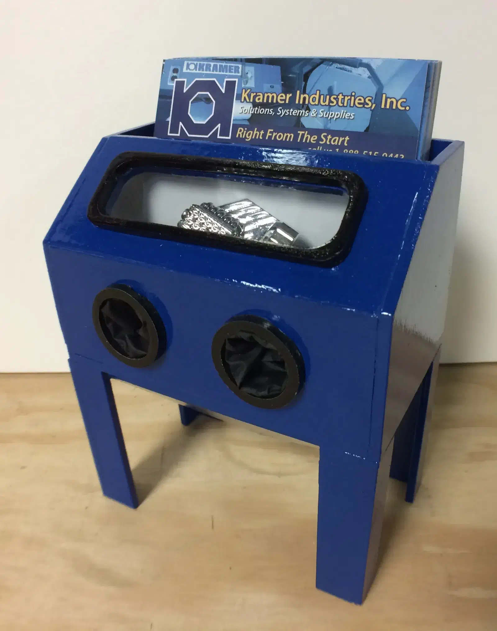 A blue miniature blasting cabinet with business cards behind its window and two small glove openings