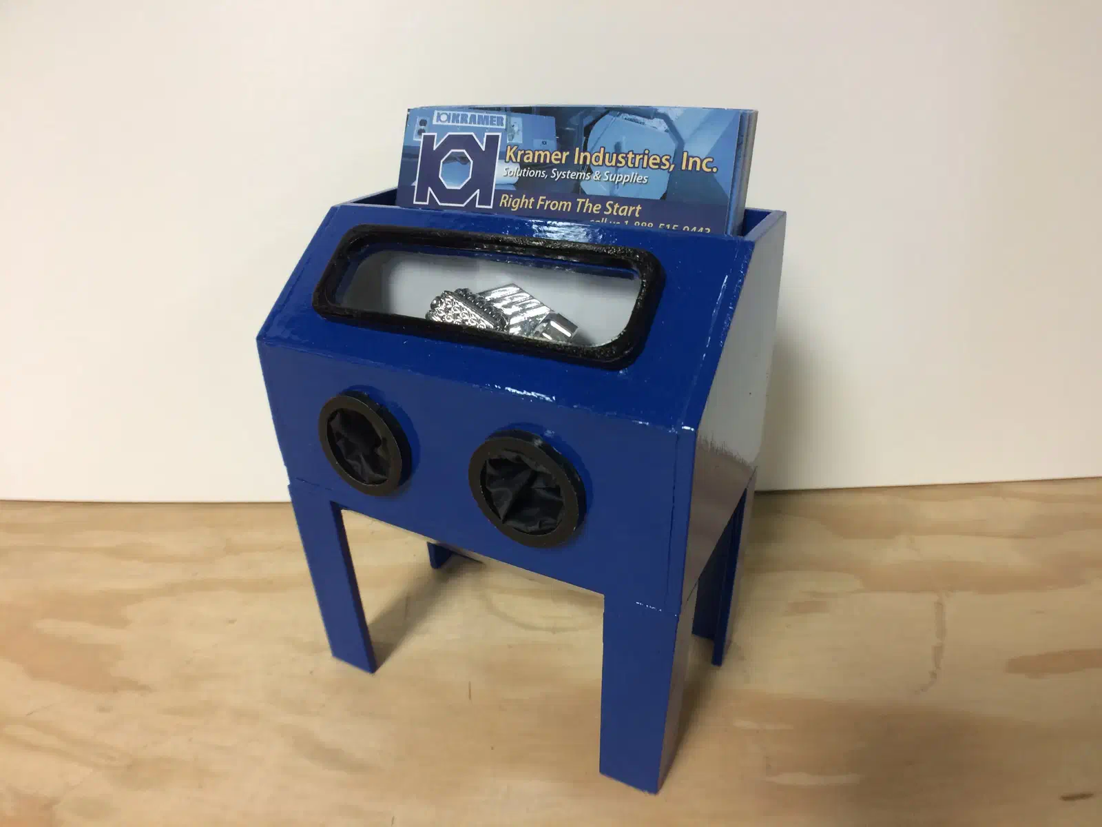 A blue miniature blasting cabinet with business cards behind its window and two small glove openings