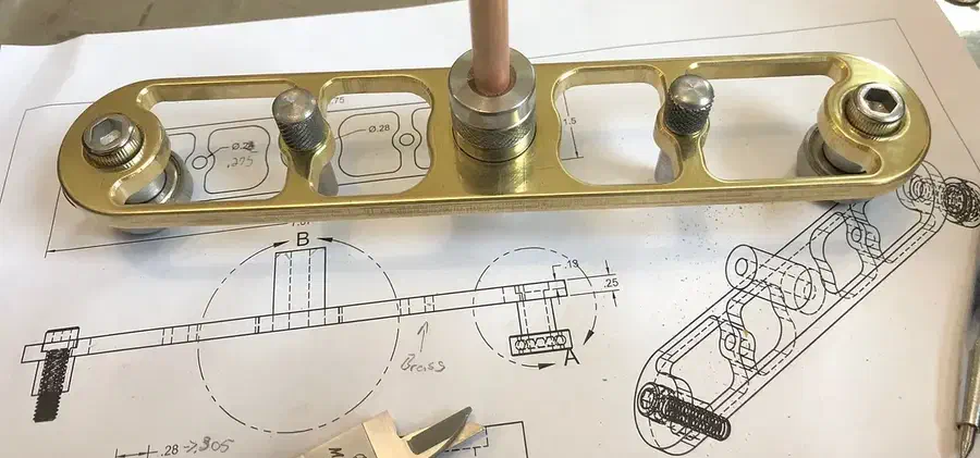 Finished brass center finder with two end pins and a pencil at its midpoint on a wooden bench