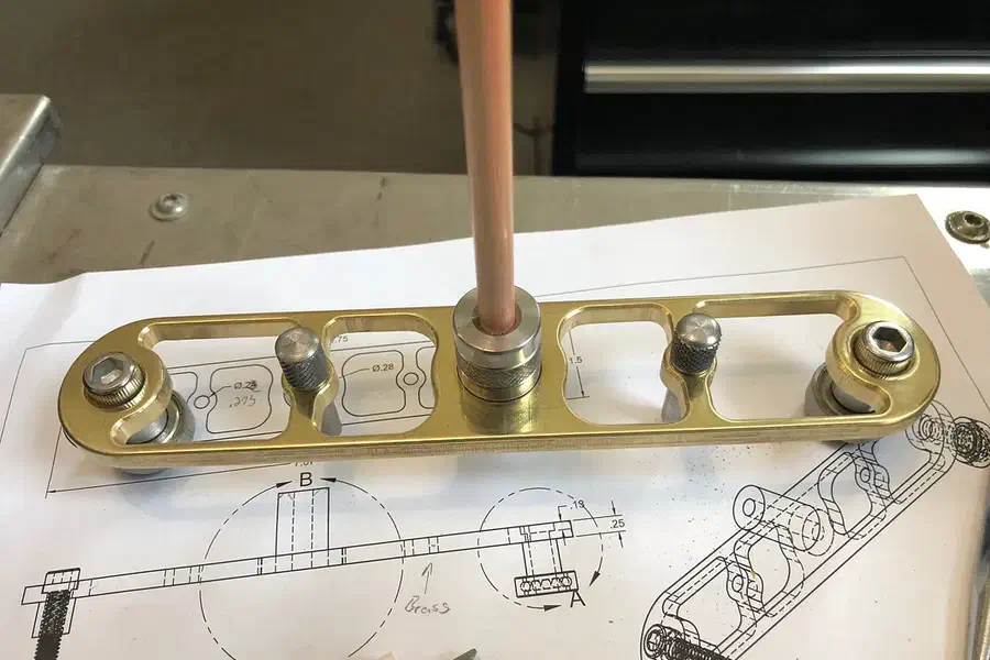 Finished brass center finder with two end pins and a pencil at its midpoint on a wooden bench