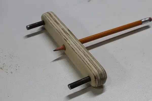 Finished layered plywood centerline tool with two metal pins and an orange pencil through its middle hole