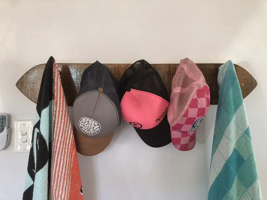 A surfboard-shaped wooden wall rack holding several caps and towels on wooden fin hooks