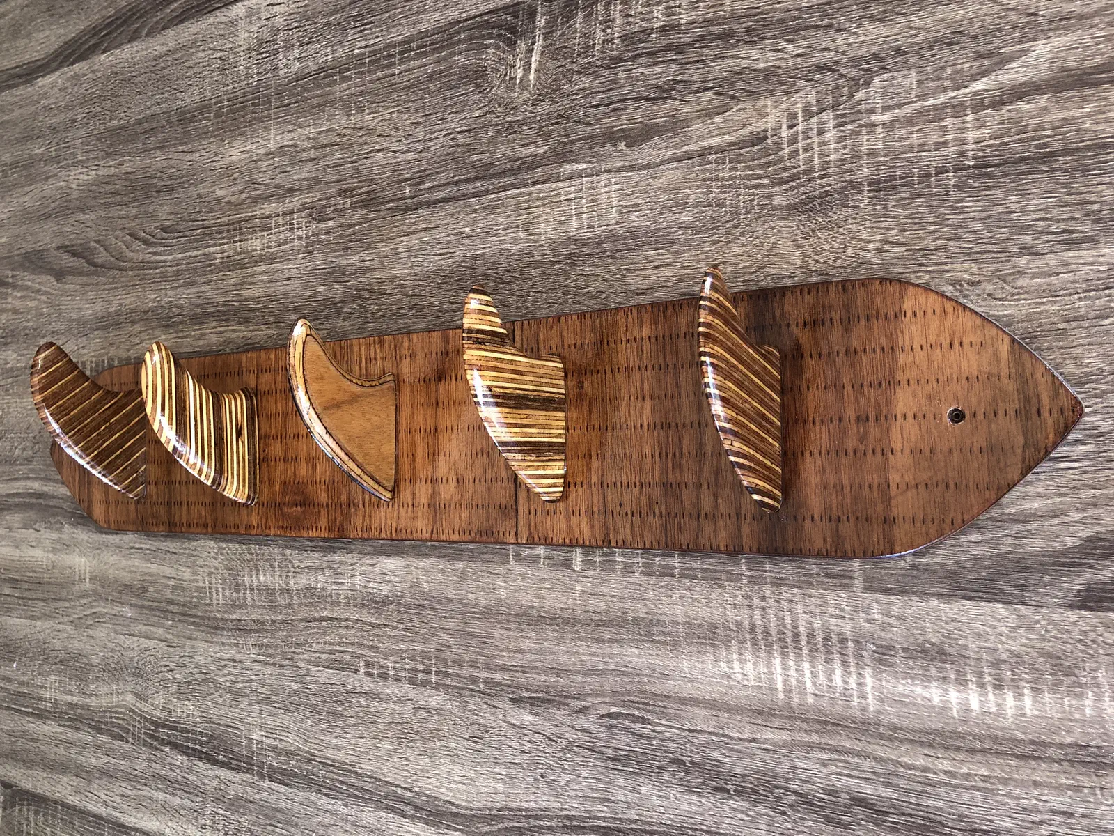 A complete wooden surfboard-shaped backboard with five striped wooden fin hooks against a gray wood-grain background