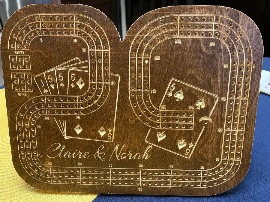 A dark wooden cribbage board shaped like the number 29, with engraved cards and the names Claire and Norah