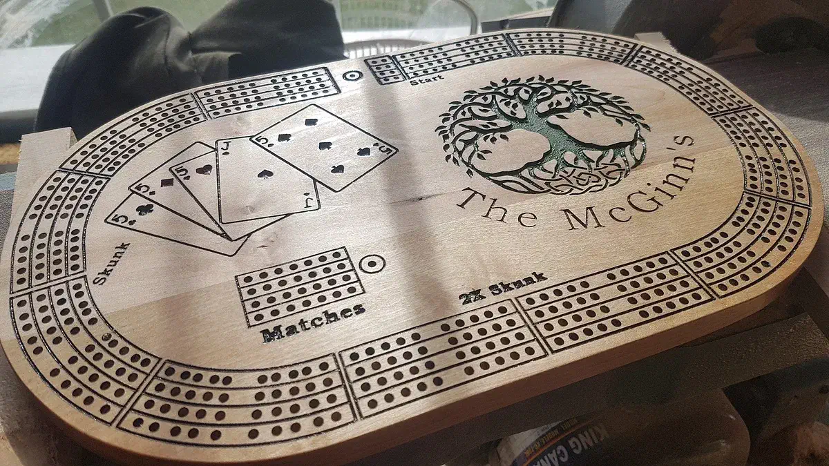 A carved oval wooden cribbage board with three peg tracks, playing cards, a green tree and the words The McGinns