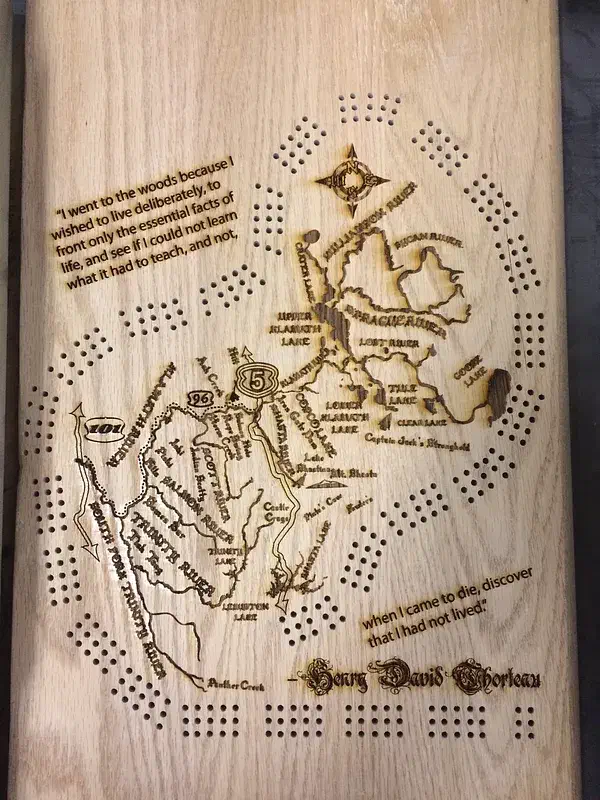 A pale wooden cribbage board with peg holes around a dark engraved map of the Klamath River watershed