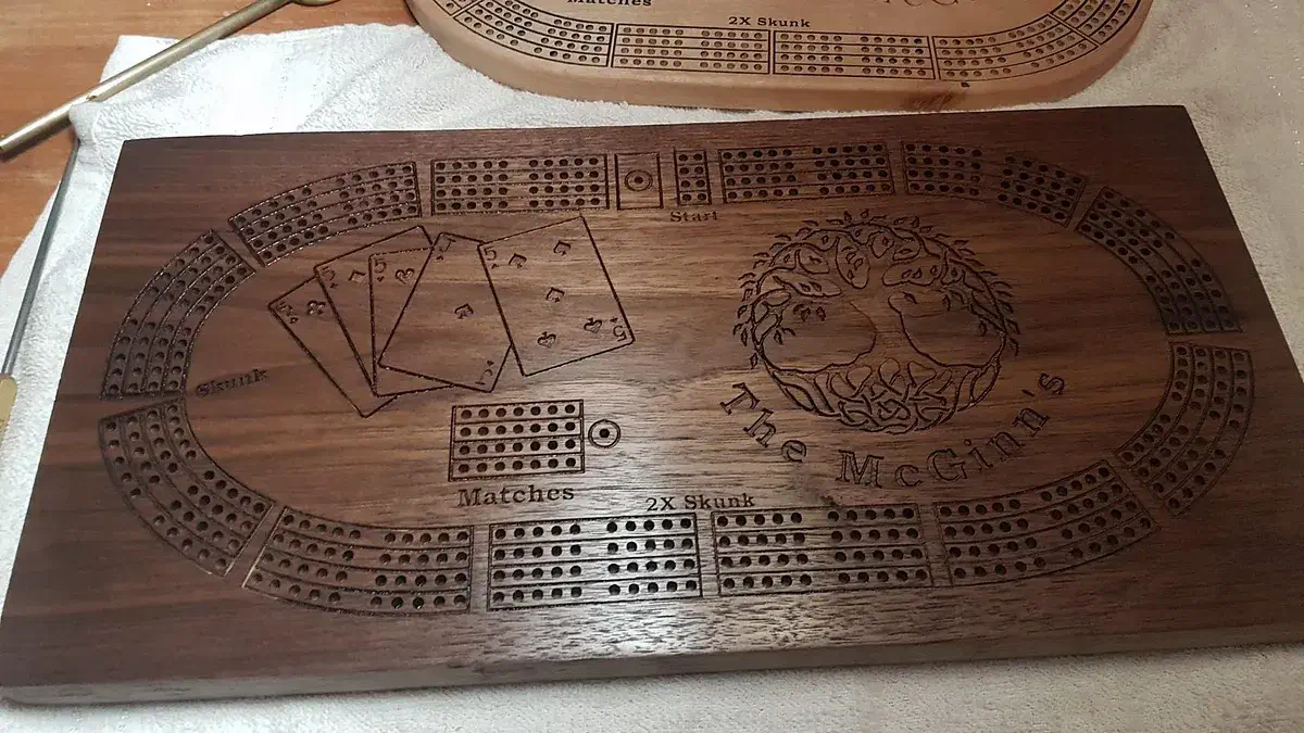 Finished wooden cribbage board with curved peg tracks, playing cards, a carved tree, and the name McGinn