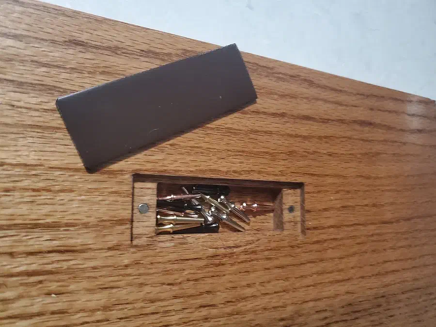 A rectangular peg pocket in a wooden board with two round magnets and a dark metal cover beside it