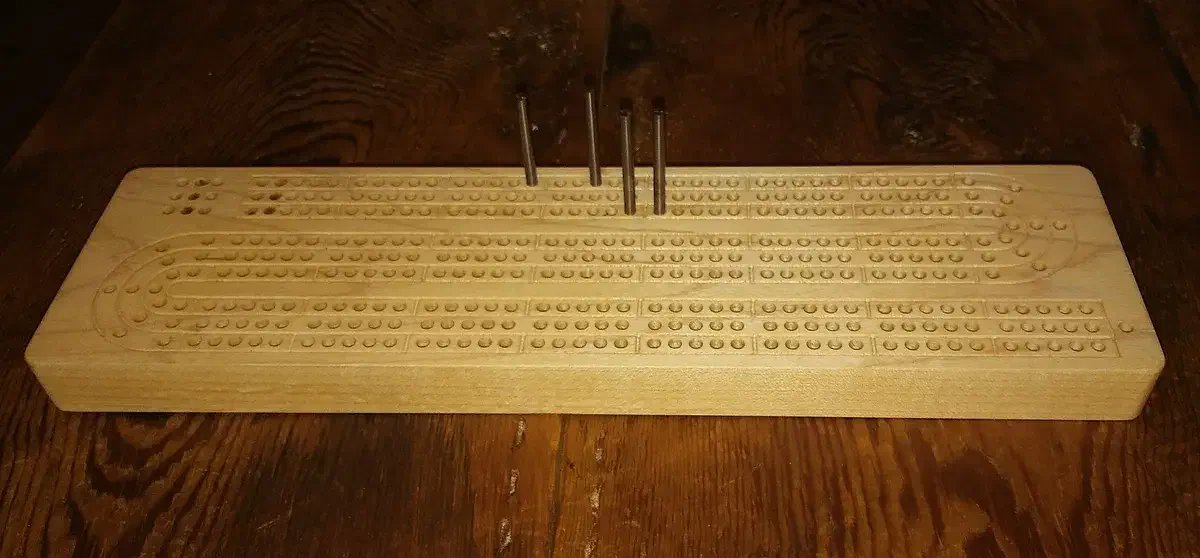 A long narrow maple cribbage board with curved scoring tracks and metal pegs standing near the center