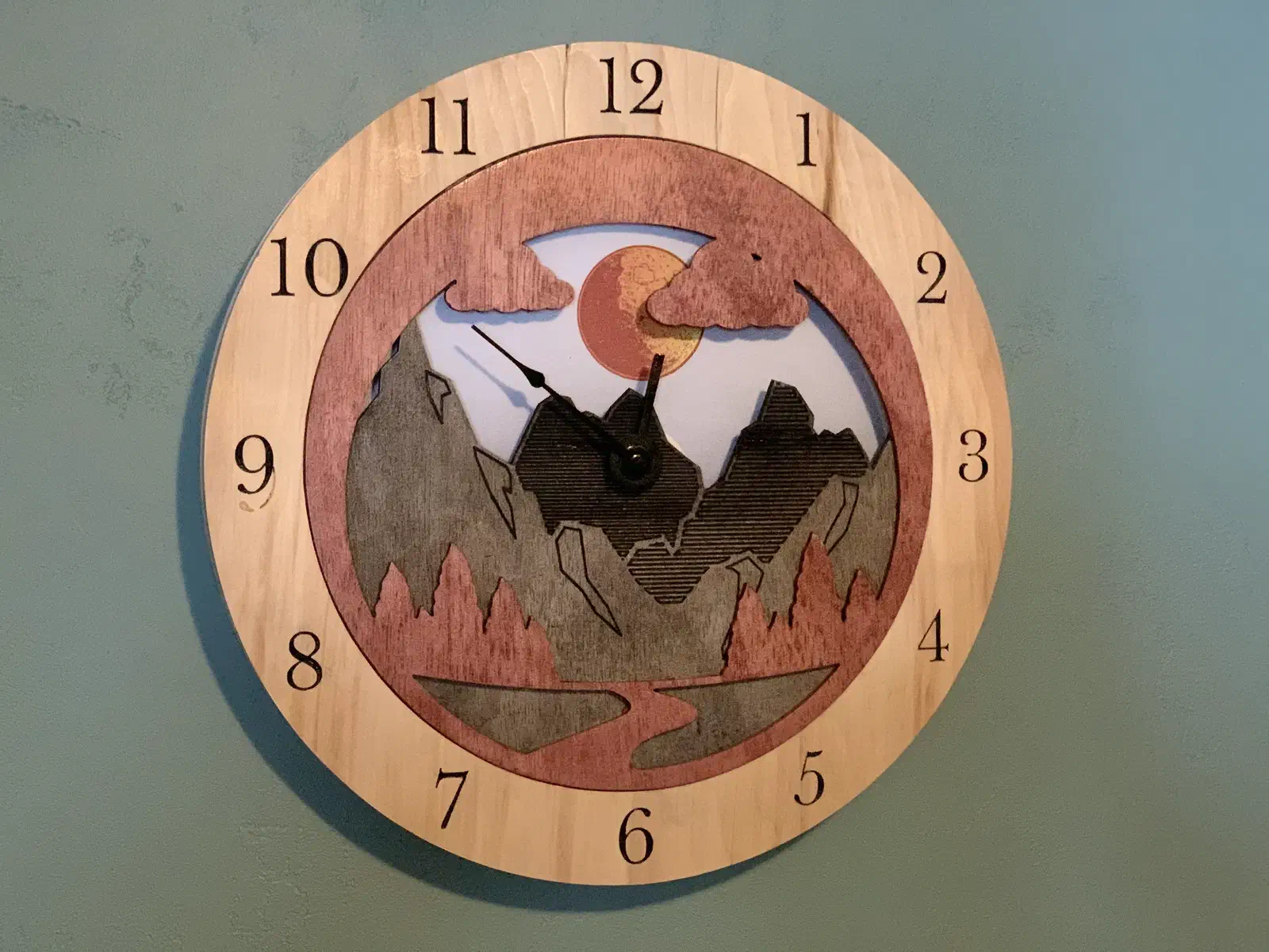 Round layered wooden clock with black numerals and a mountain landscape, clouds, sun, and river scene