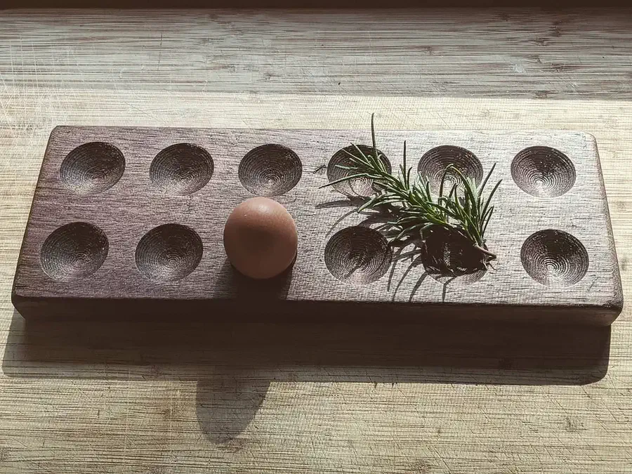 An egg and a sprig of rosemary in a rectangular wooden egg holder