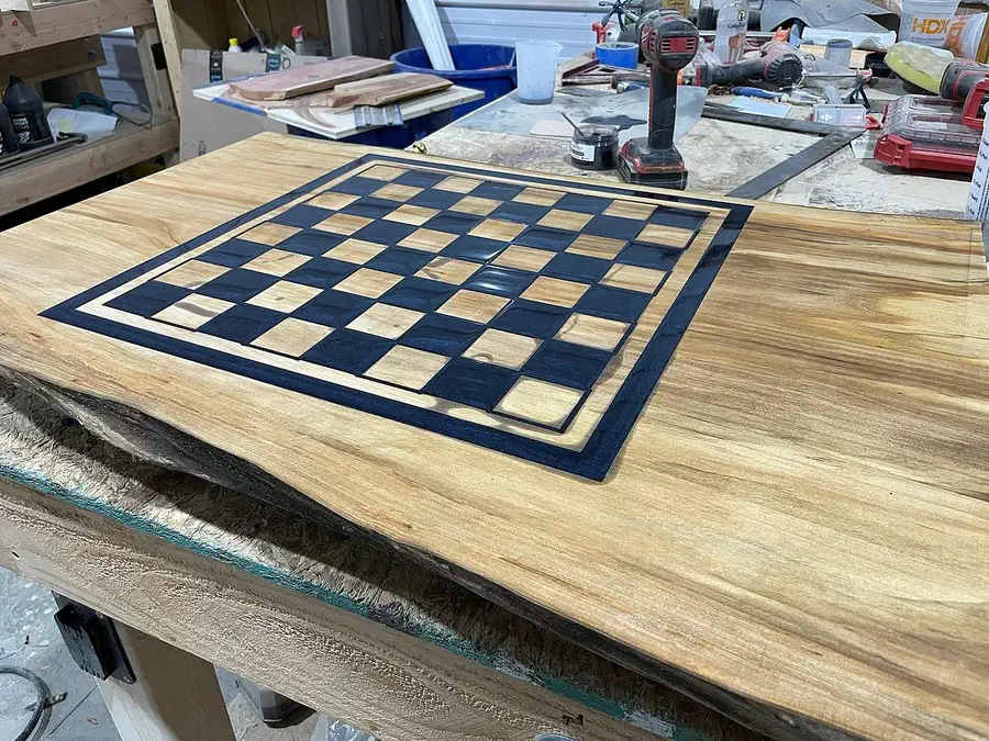A finished dark-and-light chessboard inlaid into a wide live-edge slab on a workshop bench