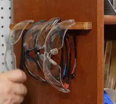 A wooden slotted rail on a cabinet holding several pairs of clear safety glasses