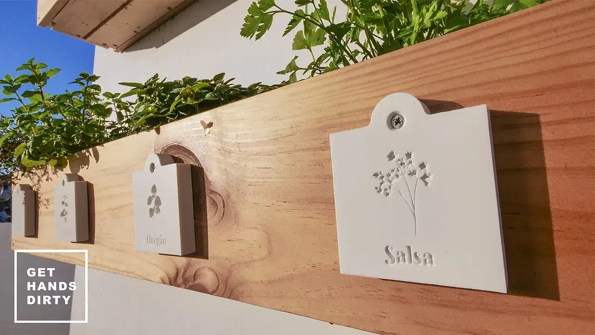 White Corian herb labels fastened to the front of a wooden planter, with an embedded Get Hands Dirty mark