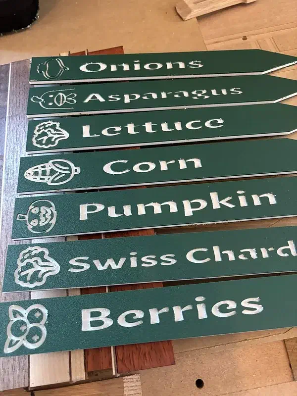 Green-and-white garden stakes with vegetable names and small vegetable drawings on a workbench