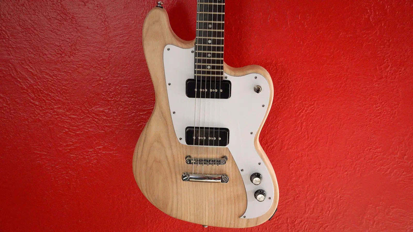 A natural-wood electric guitar body with a white pickguard, black pickups and metal bridge against a red wall