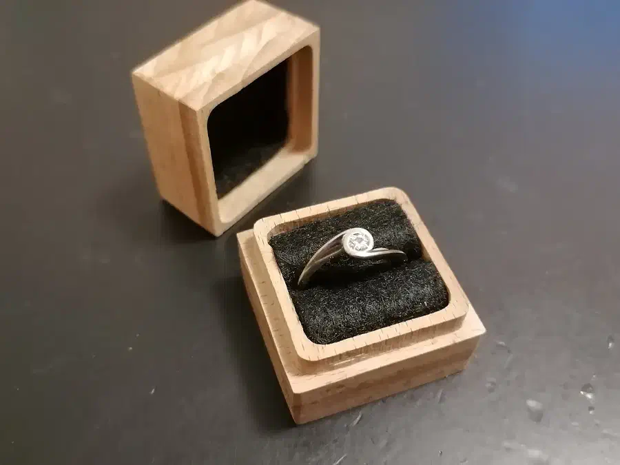Small square layered wooden box holding a ring in black felt, with its lid behind it