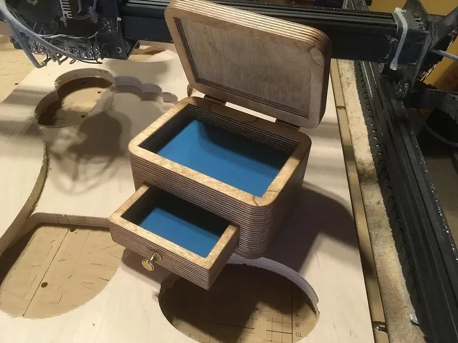 Layered plywood jewelry box with an open hinged lid, blue-lined compartment and pulled-out drawer