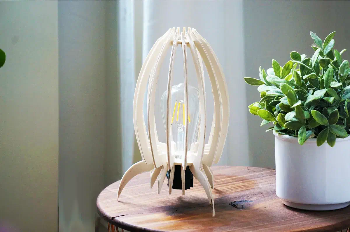 A pale plywood ribbed table lamp with pointed feet beside a potted plant