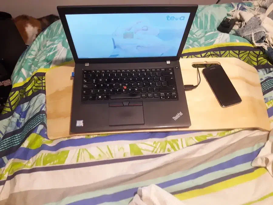 An open laptop and a phone on a pale wooden table resting on a bed