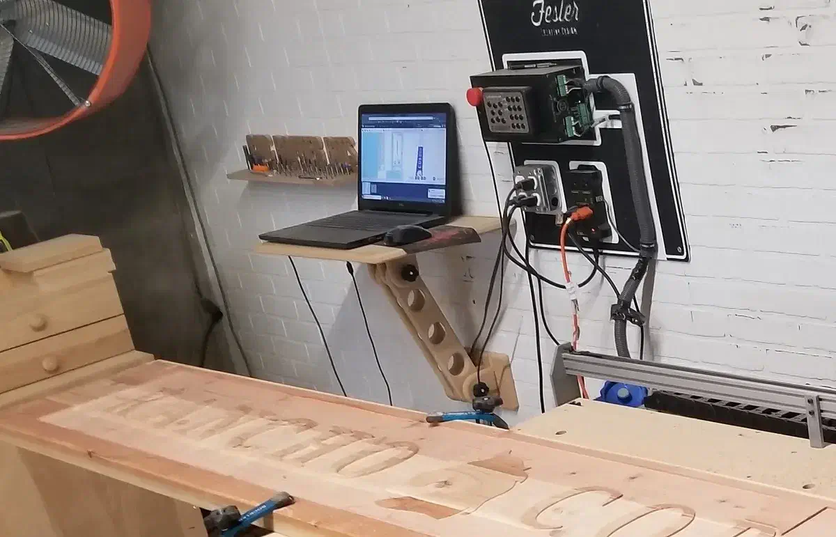 A laptop on a wooden articulating arm mounted to the wall above a CNC workspace