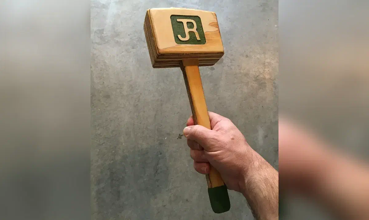 A hand holding a laminated wooden mallet with a carved R on its square head