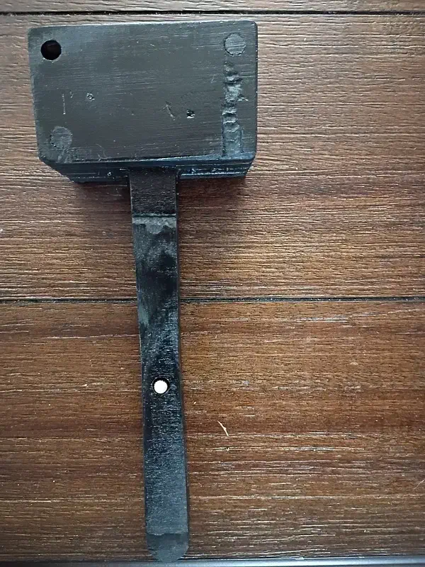 A dark-finished plywood mallet with a rectangular head, straight handle and visible round dowel ends on a wooden surface