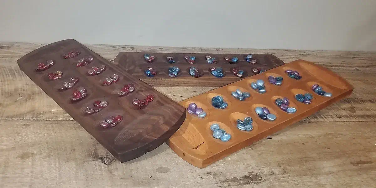 Three finished wooden Mancala boards of different shapes with red, blue, and purple glass marbles in their pits, arranged on a workbench