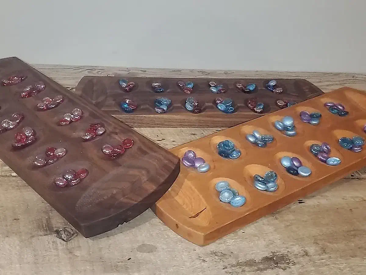 Three finished wooden Mancala boards of different shapes with red, blue, and purple glass marbles in their pits, arranged on a workbench
