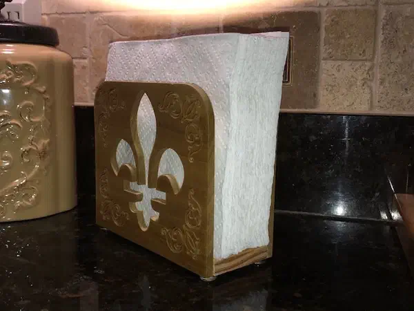 Finished poplar napkin holder with a fleur-de-lis opening and carved corner scrolls, holding paper napkins on a kitchen counter