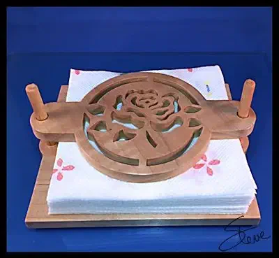 Physical wooden napkin holder with a rose-cutout top resting over a stack of napkins between two upright dowels on a blue background
