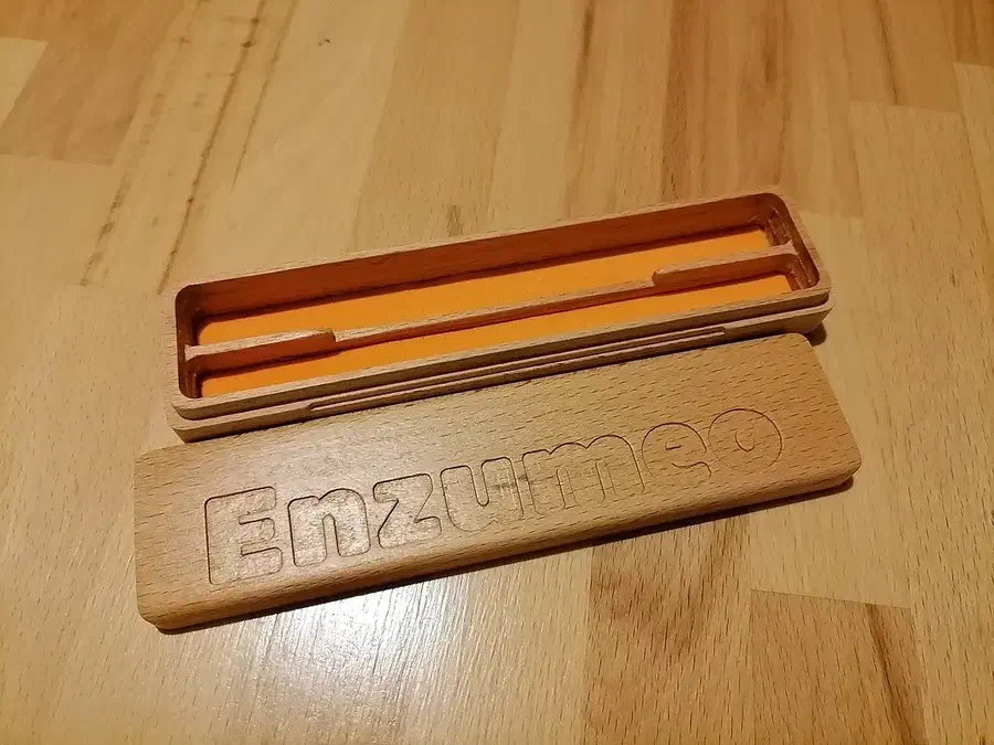 An open layered wooden pencil case with orange lining and an engraved Enzumeo lid on a wood surface