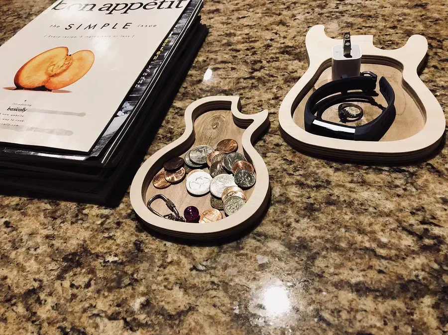 Two finished pale plywood guitar-shaped trays on a counter, one with coins and one with a watch and small charger