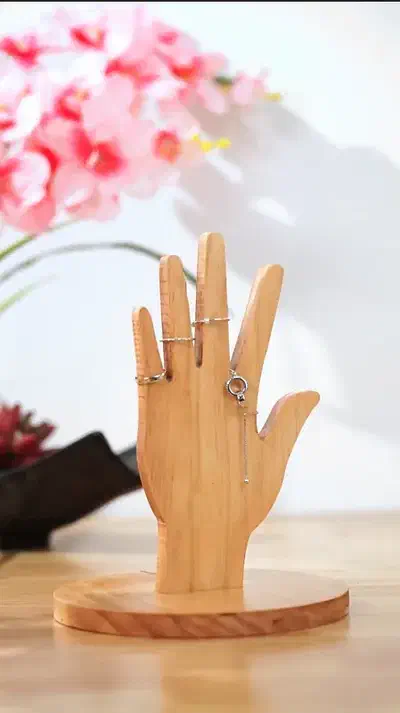 A wooden hand-shaped stand with rings on its fingers and a dangling pendant, on a round wooden base
