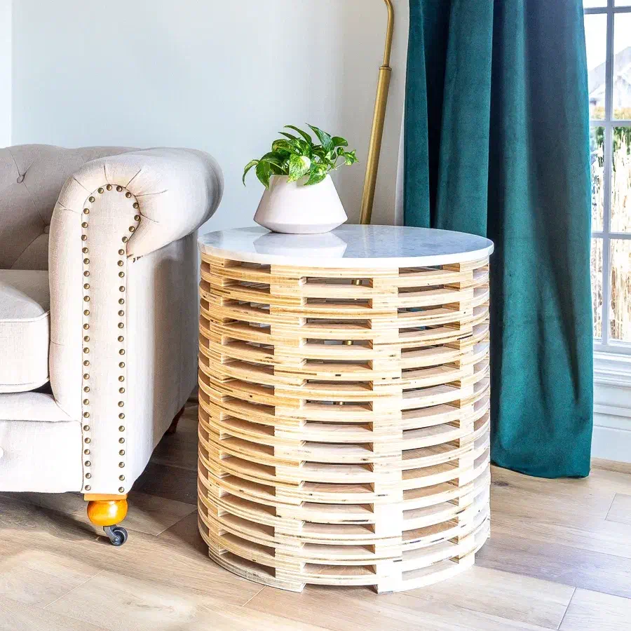 A cylindrical table built from stacked pale plywood slats with a white round top beside an upholstered armchair