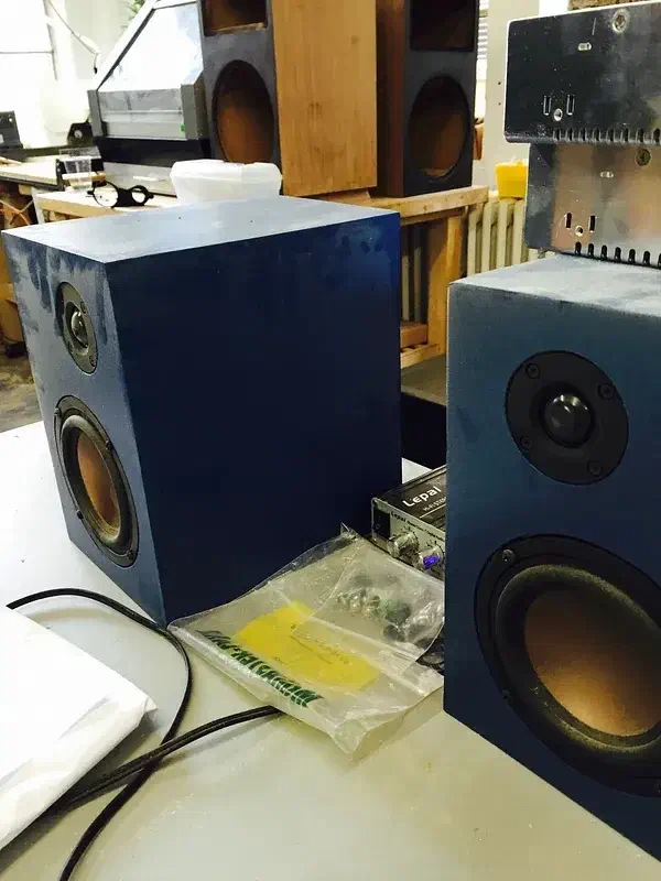 Two dark blue bookshelf speakers with small upper drivers and larger lower drivers on a workshop bench