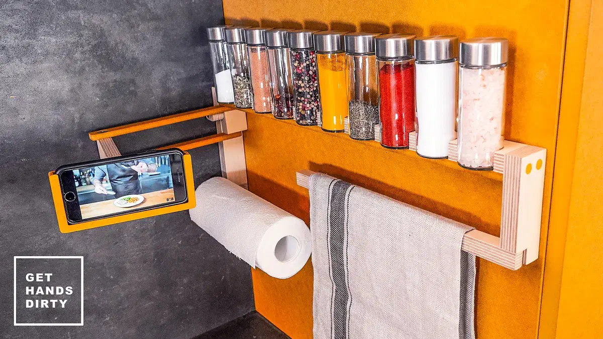 Cristiana Felgueiras’s finished wall-mounted spice shelf above a paper-towel roll, a hanging cloth and a phone support against a yellow and gray wall.