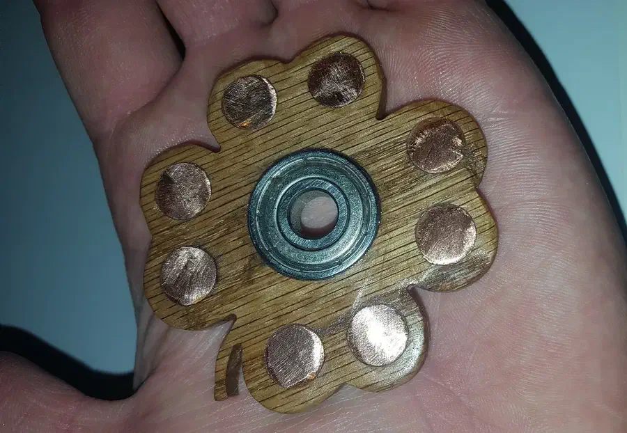 A hand holds a four-leaf wooden clover spinner with a center bearing and round copper weights