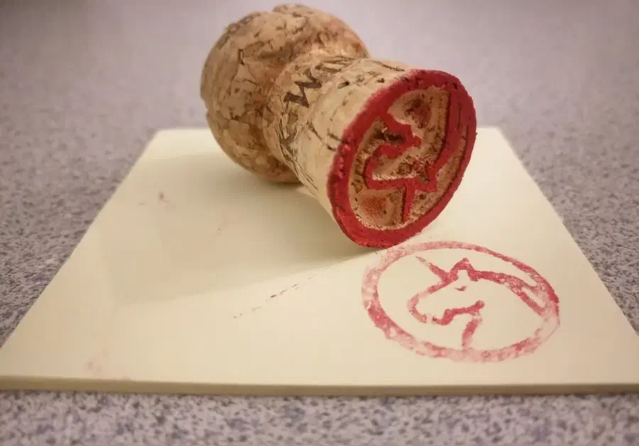A champagne cork with a red unicorn stamp face beside a small red unicorn impression on paper