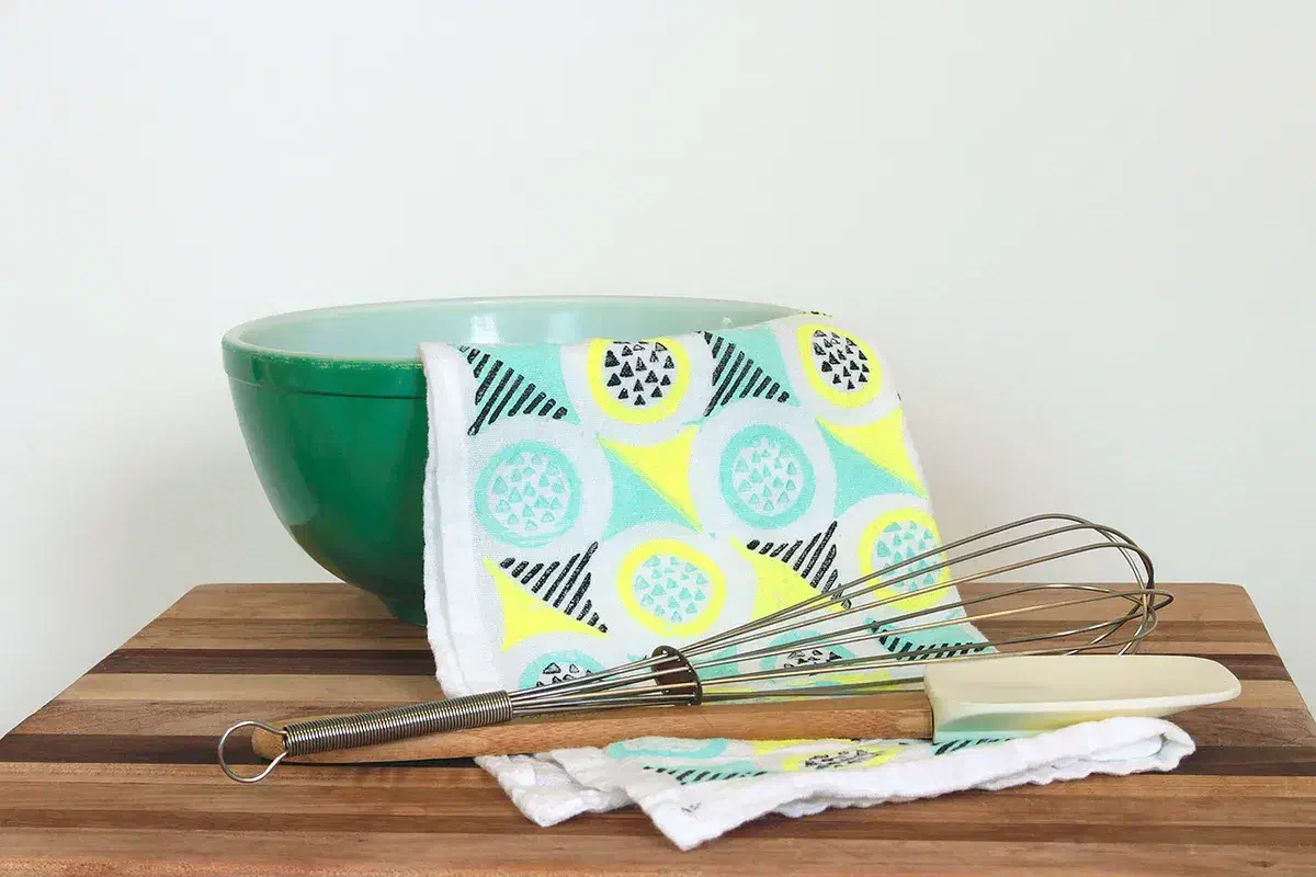 A pale tea towel with yellow, mint and black geometric block prints hanging over a mixing bowl beside kitchen utensils