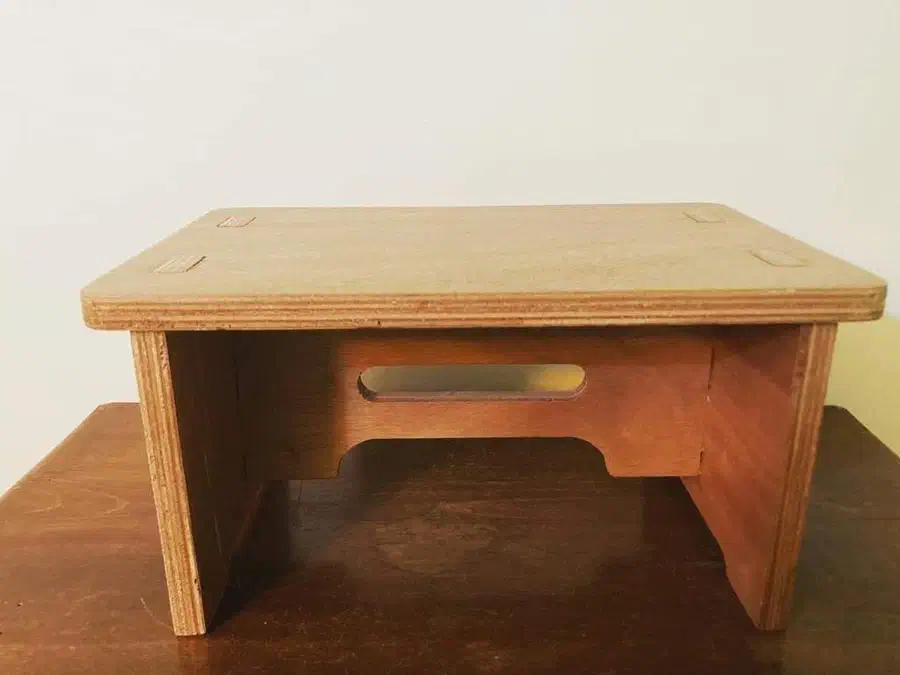 A low plywood step stool with a rectangular top, exposed plywood edges and a handhold in its cross brace
