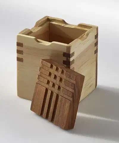 A complete light-wood box with dark corner splines and a separate patterned wooden lid leaning in front, on a plain pale background.