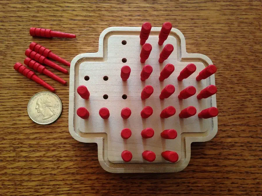 A cross-shaped wooden peg-solitaire board with red cribbage pegs and a coin beside it