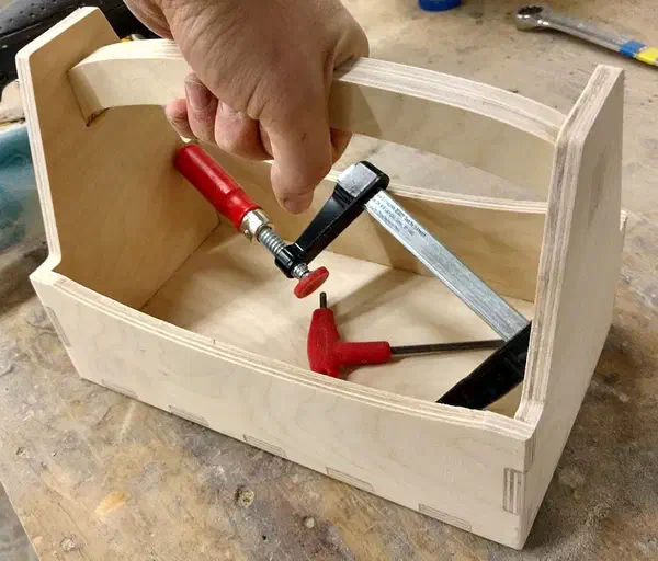 Andrew Camardella’s complete birch-plywood tote held by its curved handle, with two red-handled clamps inside on a workbench.
